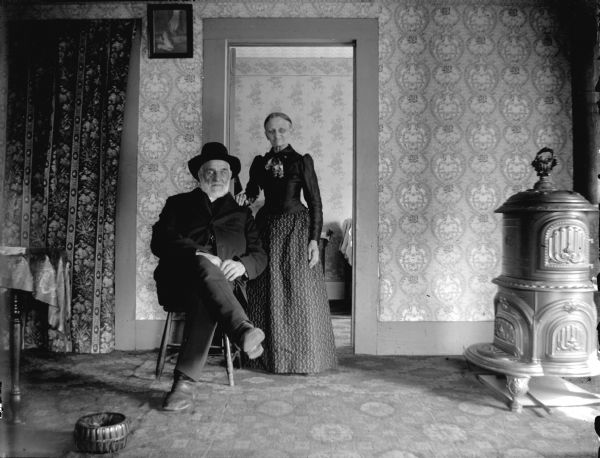 Elderly woman and man posed in a parlor with large wood stove. Probably Mr. And Mrs. Squires who owned a business in town, the Squires Oyster House.