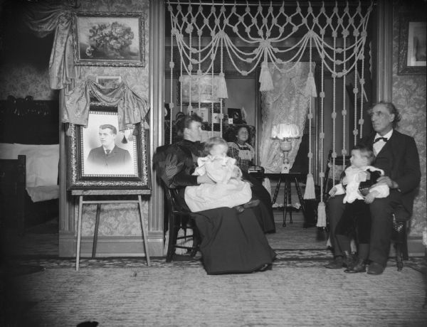 Two women, two children, and a man sitting in a parlor showing a portrait of a man and a doorway hanging with cords.
