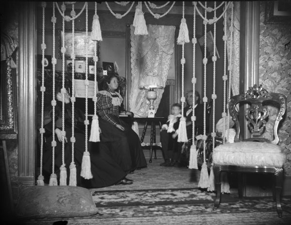 Two women, a man, and child sitting in a parlor with a doorway hanging with decorative cords.