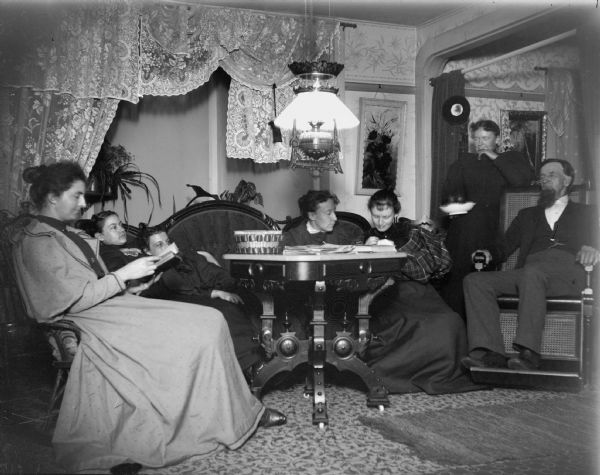 Six women and a man gathered at a parlor table at a boarding house. The man is probably Amos Elliott, and the women around the table are teachers studying for the evening.