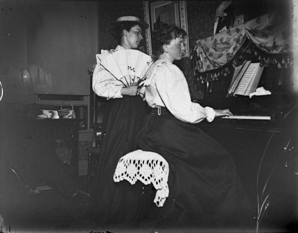Woman posed playing a piano while another woman stands and holds a folding fan.
