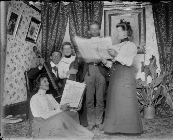 Three women and two men playing instruments and singing in a parlor. Man plays a bass, while a woman plays a guitar.