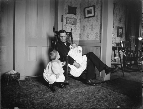 Man sits in a rocking chair holding a baby, while a child sits on a stool next to him. The man is possibly John H. Mills, holding Dudley Mills, and the child is possibly Philip B. Mills.	