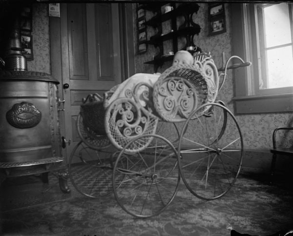 Infant in a high-wheeled baby carriage next to a wood stove.