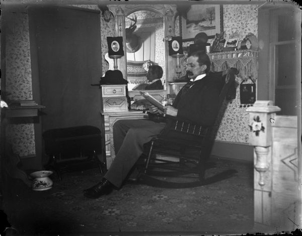 Man seated in a rocking chair reading with deer head mount reflected in mirror.