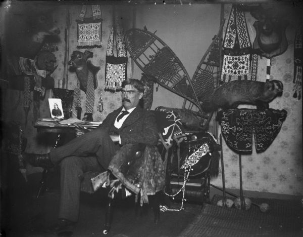 Man seated in front of a wall and table displaying snowshoes, stuffed animals, and Native American artifacts, probably Tom Roddy.