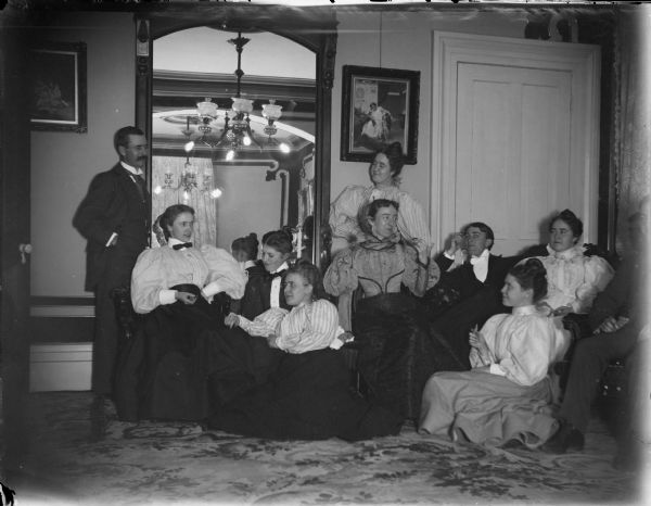 Large group of seven women and three men, all in evening dress, conversing in a parlor in front of a large mirror. Probably the Spaulding house and some Spaulding family members, including the twins.