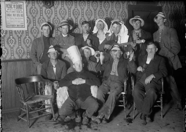 Group of a dozen men mugging for the camera. Poster on the wall advertises a "Grand Picnic and Dance in Miller's Bowery, Caroline, Wis., Sunday, May 18th 1913."