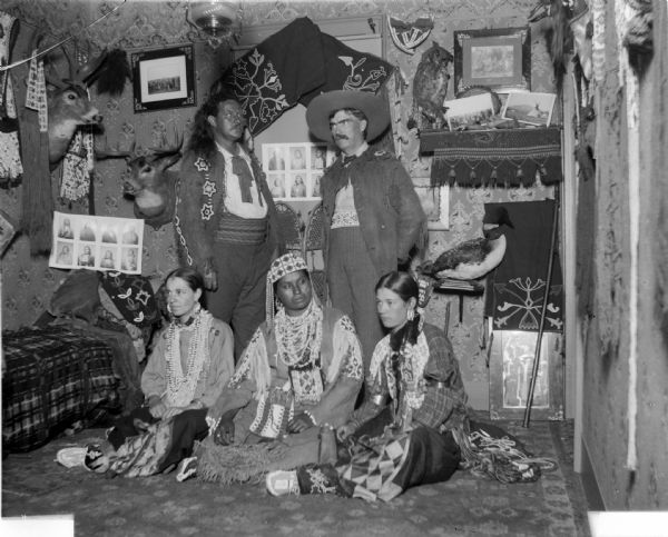Two white women and three white men pose as Native Americans in a home with numerous artifacts including several taxidermy specimens.