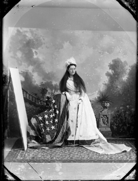 Studio portrait of a woman with long hair and a trailing dress leaning on a United States flag in front of a painted backdrop.
