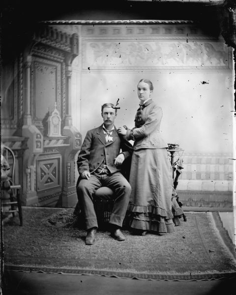 Studio portrait of a man sitting and a woman standing in front of a painted backdrop.