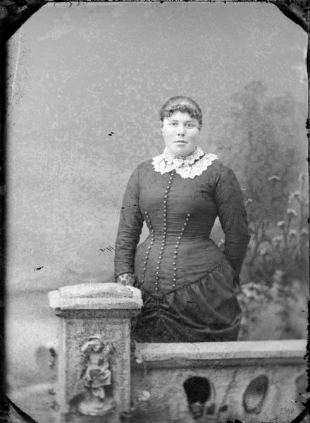 Studio portrait of a woman standing behind a stone fence in front of a painted backdrop.