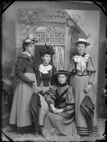Studio portrait of four women in hats, two standing and two sitting, in front of a painted backdrop.