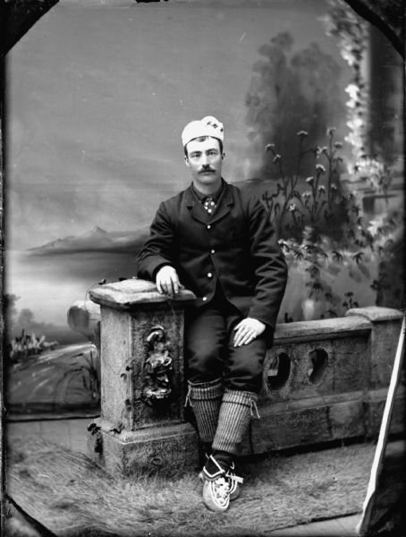 Studio portrait of a man sitting on a stone fence in front of a painted backdrop, probably dressed in a Norwegian costume.