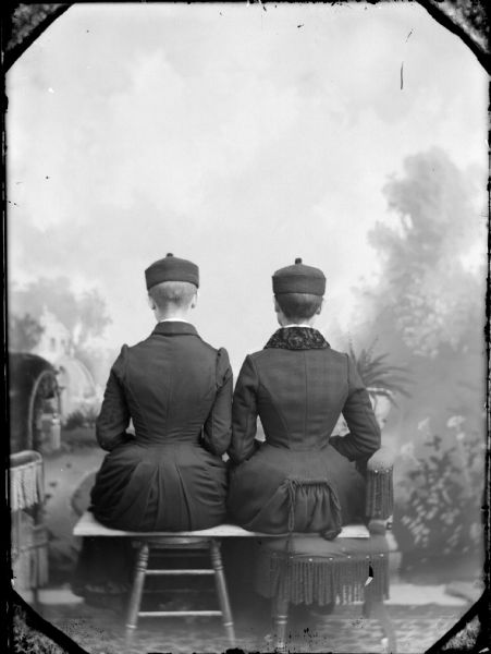 Studio portrait of two women with short hair sitting with their backs to the camera in front of a painted backdrop.