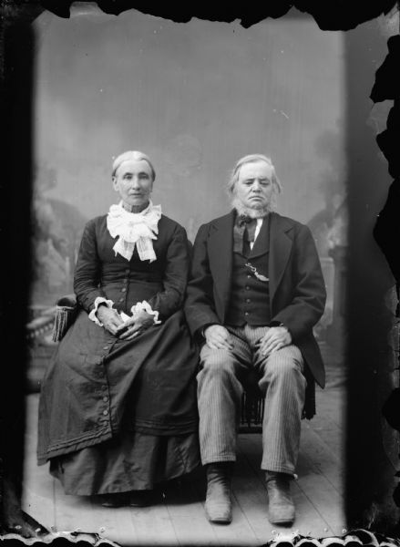 Studio portrait of an elderly man and woman sitting in front of a painted backdrop. Identified as Eli and Mary Tiffany of Melrose by their descendant Jeff Rand of La Crosse, Wisconsin, in 1986 from photographs with over-painting in the family's possession.