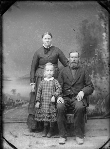 Studio portrait of a man sitting, and a woman and girl standing, in front of a painted backdrop.