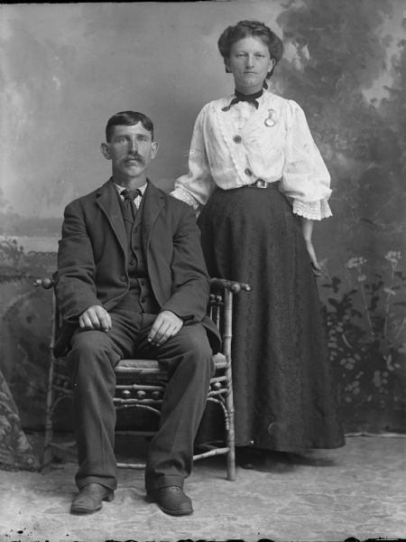 Studio portrait of a seated man and standing woman in front of a painted backdrop. The woman is wearing a long skirt with a puffy blouse, and the man is wearing a suit jacket, necktie, vest and trousers.