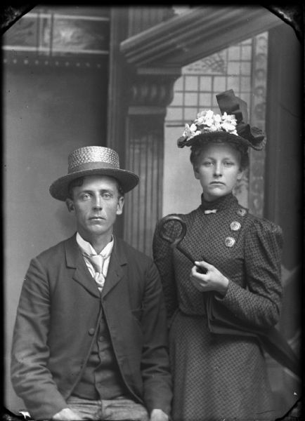 Studio portrait of a seated man and a standing woman holding an umbrella in front of a painted backdrop. Both are wearing hats. The man is wearing a suit jacket, vest, trousers and necktie.