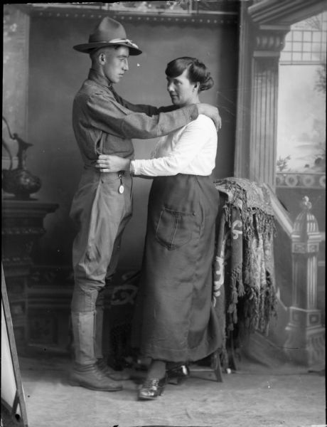 Studio portrait of a standing soldier with his arms on the shoulders of a woman who is standing and who has her hands on his waist. They are posing in front of a painted backdrop.