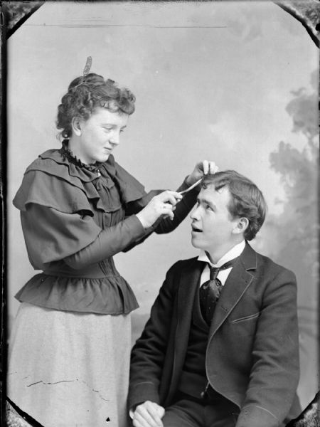 Studio portrait of a standing woman curling the hair of a seated man in front of a painted backdrop.