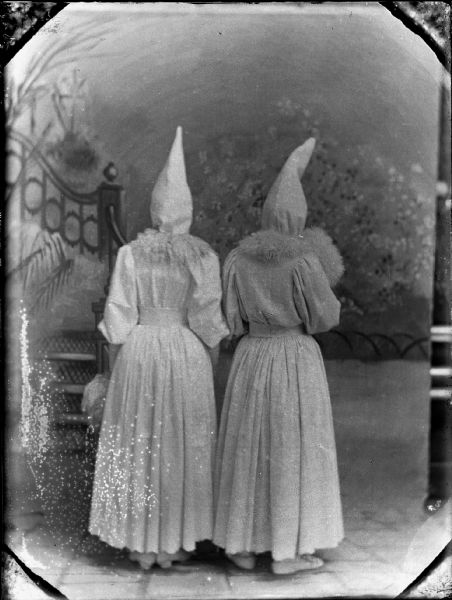 Studio portrait of two women wearing white pointed hats and long, white dresses with fluffy collars, and broad white belts. They stand their backs to the camera before a painted studio background which depicts a staircase leading to left. They are possibly actors dressed for a play.