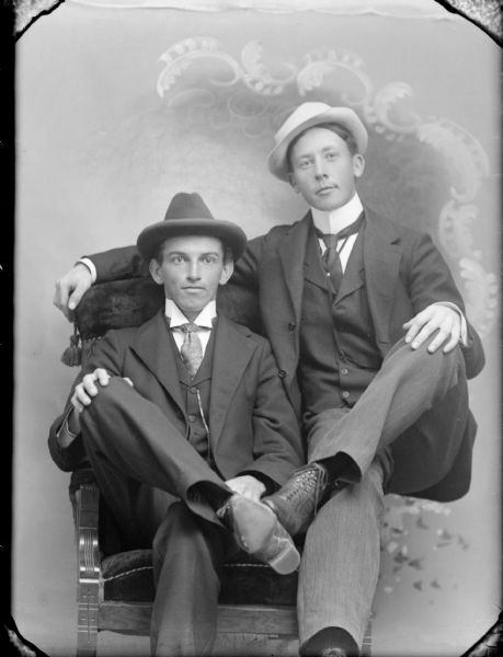 Studio portrait of two seated men with their legs crossed toward each other. One man has his arm over the chair back, and is sitting on the arm of the chair.  Both wear suits with vests, trousers and neckties, with high collared shirts. They are both wearing hats and are posing in front of a painted backdrop.
