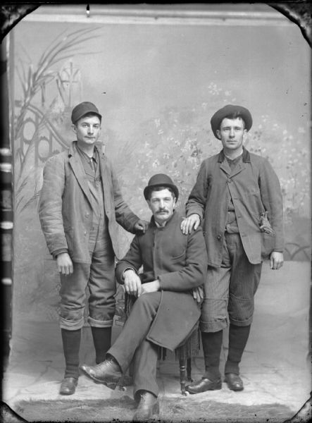 Studio portrait of three men wearing hats, posing in front of a painted backdrop, who are standing with each of their hands on the shoulders of a seated man, who has his legs crossed.