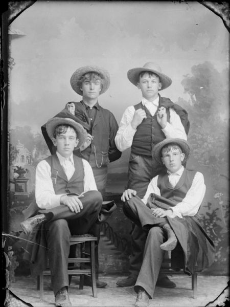 Studio portrait of four boys in hats posing in front of a painted backdrop. They are all wearing vests, and are holding or carrying their suit jackets. Two are standing and the other two are sitting.