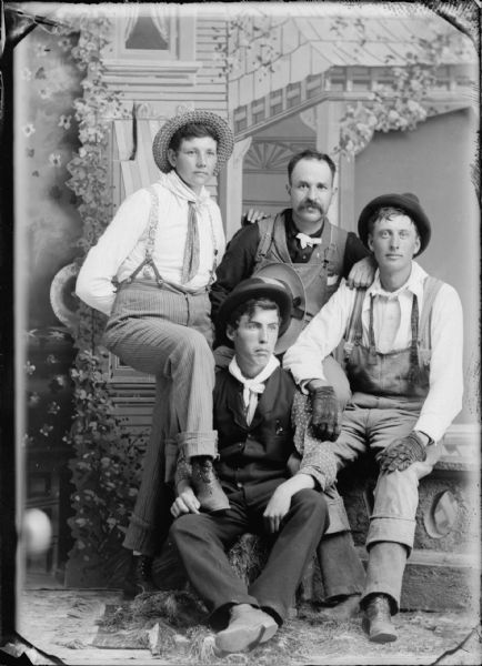 Studio portrait of three seated men dressed in work clothes, and a standing woman wearing trousers and suspenders. They are posing in front of a painted backdrop. Three are wearing hats, one is holding a hat, and one man wears gloves.