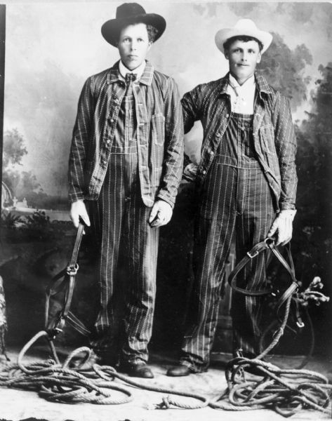 Studio portrait of two men posed standing, wearing matching striped overalls with matching jackets. They are holding bridles attached to ropes and are posing in front of a painted backdrop.