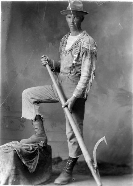 Studio portrait of a lumberjack wearing a hat, fringed shirt, trousers, suspenders, and what appear to be spiked boots. He is holding a peavey and is posing with one foot up on a box in front of a painted backdrop.