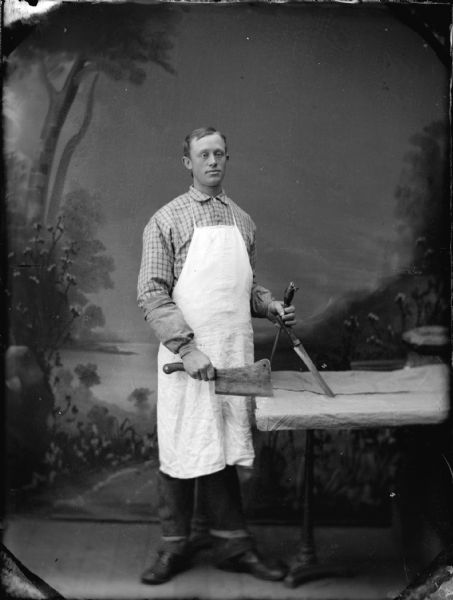 Studio portrait in front of a painted backdrop of a butcher wearing an apron, and holding a cleaver and saw.