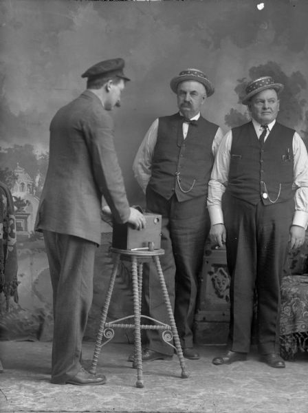Studio portrait of three standing men, two wearing matching straw hats, and vests with trousers, and the other holding a camera on a stool. They are posing in front of a painted backdrop. From right to left, Olaf Olson, Jack Taylor, and an unidentified traveling pottery salesman.