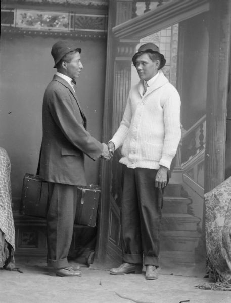 Studio portrait of two standing men shaking hands posing in front of a painted backdrop, probably Ho-Chunk Indians. Both men are wearing hats, and the man on the left is wearing a suit jacket and trousers, the man on the right wears a sweater over trousers.