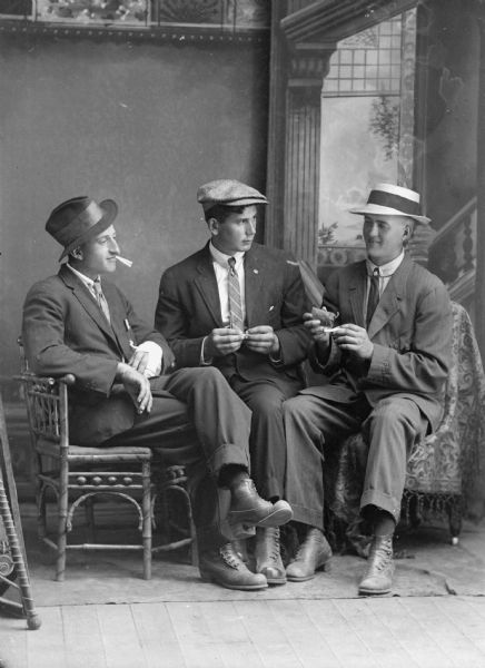 Studio portrait of three seated men making cigarettes, posing in front of a painted backdrop. They are all wearing suit jackets, neckties, and hats. Probably Phil Nutter in the center and Earl Davis to the right.