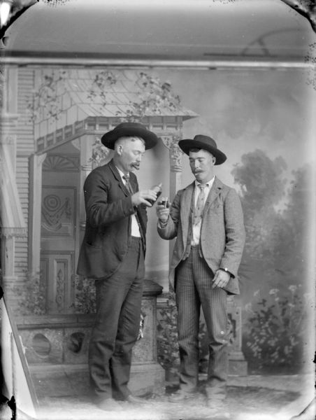 Studio portrait of two standing man smoking cigars posing in front of a painted backdrop. One man is pouring liquid into a glass. Both men are wearing hats, suit jackets, vests, and trousers.