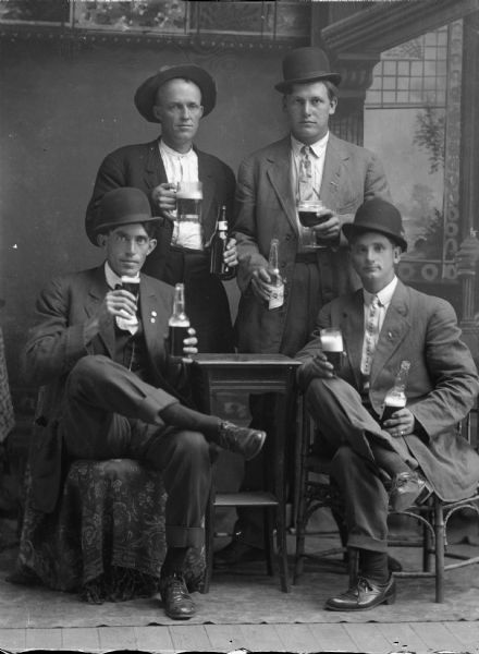 Studio portrait of four men in hats, suit jackets, vests, and trousers. Two men are standing and two sitting around a small table, drinking from glasses filled from beer bottles posing in front of a painted backdrop. Man standing on the left is probably John Tester, a Spanish-American War veteran.