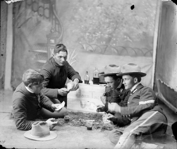 Studio portrait of four soldiers lying on the ground by a box that says, "Hard Tack U.S.," playing cards and drinking. They are posing in front of a painted backdrop.