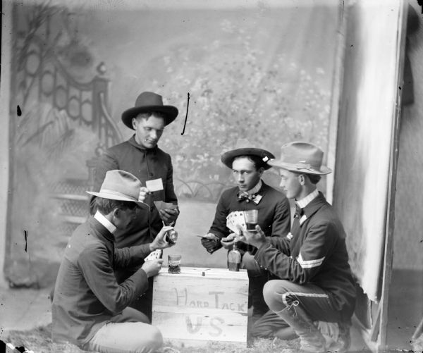 Studio portrait of four soldiers kneeling around a box that says, "Hard Tack U.S.," playing cards and drinking. They are posing in front of a painted backdrop.