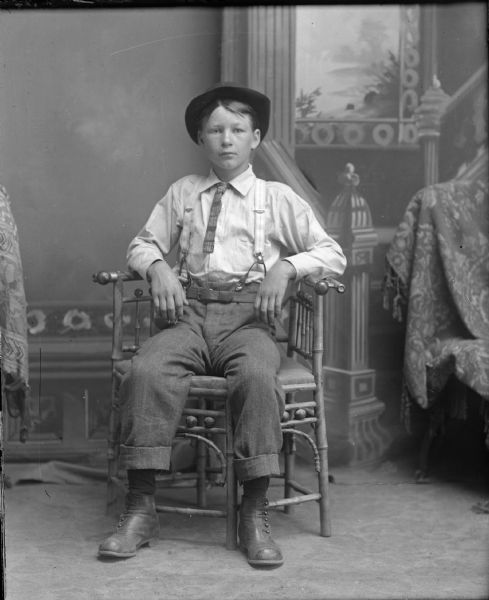 Studio portrait of a seated boy in a hat and necktie in front of a painted backdrop.