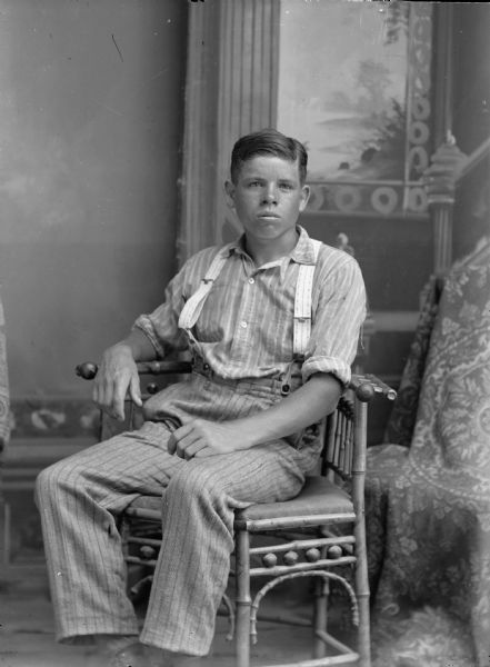 Studio portrait of a seated young man wearing a striped shirt, suspenders, and trousers in front of a painted backdrop.