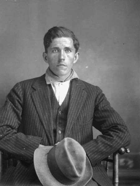 Studio portrait of a seated young man in a striped suit jacket and vest holding his hat in hand. He wears a collarless shirt with fabric tucked around his neck. He sits in a spindle chair in front of a plain painted backdrop.