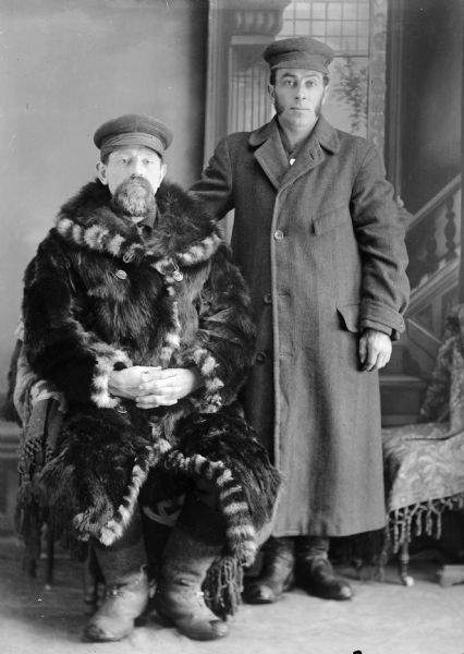 Studio portrait of two men in winter coats posing in front of a painted backdrop. One is in a wool coat, the other in a fur coat.
