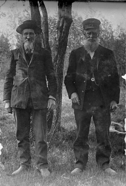 Two elderly men in front of young tree, both with beards and moustaches, wearing outer jackets. The Man on the right wears an engineer's cap, and a watch chain in his vest. The man on the left wears a tailored jacket and hat with upturned brim.
