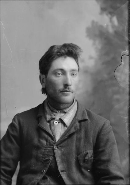 Studio portrait in front of a painted backdrop of a seated young man looking off to the right. His tabbed jacket is patched just below the lapel, on left side, and his pocket on right is disheveled. His jacket, buttoned at the top button only, covers  two shirts, the innermost is collarless. He sports a short early moustache, and hair below his ears, short along the sides.