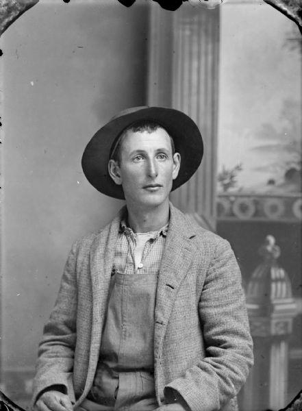 Studio portrait of a man wearing a suit coat over overalls, work shirt, and undershirt with a fedora hat. He is seated before a painted backdrop depicting an intricate newel post and window. He is probably either Hank or Fred Bahnub.