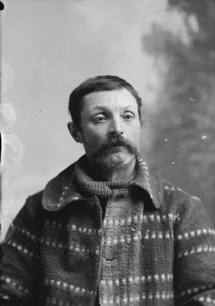 Studio portrait of a seated man with a moustache in a knit sweater. Probably Alex Bock.