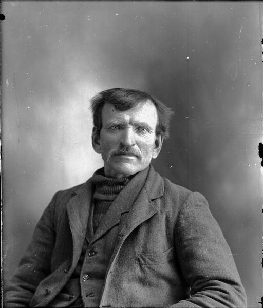 Studio portrait of a seated man with a moustache in a sweater, vest, and suit coat.