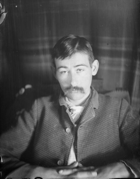 Portrait of a seated man with moustache wearing a suit jacket with a white necktie.